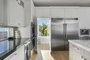 Kitchen with white cabinetry, appliances with stainless steel finishes, light wood-type flooring, dark stone countertops, and beverage cooler