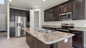 Kitchen with dark brown cabinetry, appliances with stainless steel finishes, sink, light tile patterned floors, and a center island with sink