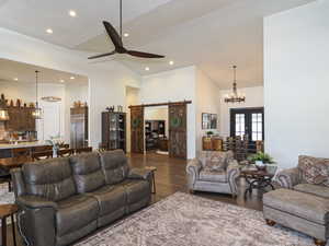 Living room featuring a barn door, dark wood-type flooring, high vaulted ceiling, french doors, and ceiling fan with notable chandelier