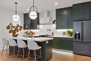 Kitchen with a center island with sink, pendant lighting, built in appliances, light hardwood / wood-style flooring, and wall chimney exhaust hood