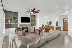 Tiled living room with ceiling fan with notable chandelier, a tile fireplace, and built in features