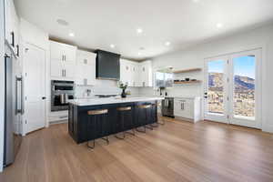 Kitchen with wall chimney range hood, light hardwood / wood-style floors, a center island, appliances with stainless steel finishes, and white cabinets