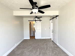 Unfurnished bedroom featuring a textured ceiling, ceiling fan, carpet flooring, and a barn door