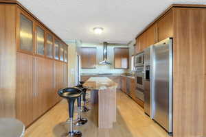 Kitchen featuring stainless steel appliances, a kitchen breakfast bar, light wood-type flooring, a kitchen island, and wall chimney exhaust hood