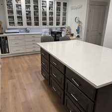 Kitchen with dark brown cabinetry, white cabinets, light wood-type flooring, light stone countertops, and glass insert cabinets