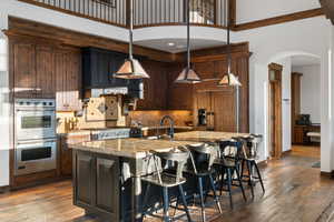 Kitchen featuring a kitchen island with sink, a towering ceiling, hanging light fixtures, double oven, and light stone counters