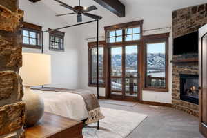 Carpeted bedroom with ceiling fan, access to exterior, beam ceiling, multiple windows, and a stone fireplace