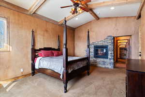 Carpeted bedroom with ceiling fan, lofted ceiling with beams, and a stone fireplace