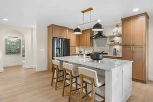 Kitchen with a center island with sink, high end fridge, backsplash, wall chimney range hood, and light stone counters