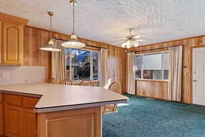 Kitchen with a textured ceiling, wooden walls, hanging light fixtures, ceiling fan, and dark carpet