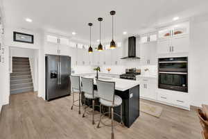 Kitchen with white cabinetry, wall chimney range hood, pendant lighting, stainless steel appliances, and a center island with sink