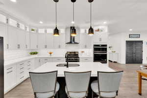 Kitchen featuring white cabinetry, hanging light fixtures, a kitchen island with sink, sink, and wall chimney exhaust hood