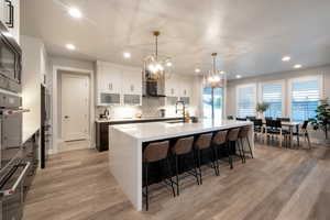 Kitchen featuring white cabinetry, sink, pendant lighting, an island with sink, and wall chimney exhaust hood