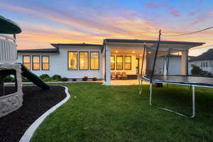 Back house at dusk featuring a trampoline, a lawn, and a patio area