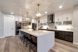 Kitchen with appliances with stainless steel finishes, wall chimney range hood, decorative light fixtures, white cabinets, and a center island with sink