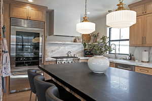 Kitchen featuring a kitchen bar, appliances with stainless steel finishes, tasteful backsplash, hanging light fixtures, and wall chimney exhaust hood