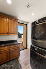 Clothes washing area with stacked washer and dryer, a textured ceiling, cabinets, and light hardwood / wood-style flooring