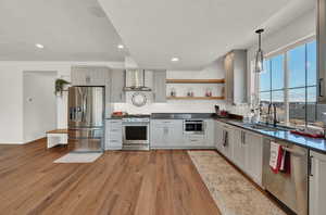 Kitchen with light hardwood / wood-style flooring, appliances with stainless steel finishes, sink, wall chimney exhaust hood, and decorative light fixtures