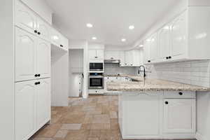 Kitchen with oven, light stone countertops, white cabinetry, sink, and backsplash