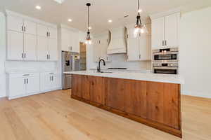 Kitchen featuring white cabinets, stainless steel appliances, premium range hood, hanging light fixtures, and a center island with sink