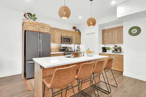 Kitchen featuring a center island with sink, stainless steel appliances, light brown cabinets, dark wood-type flooring, and pendant lighting