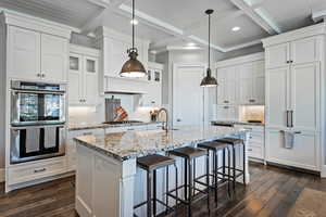 Kitchen featuring white cabinets, stainless steel appliances, beamed ceiling, an island with sink, and light stone counters