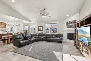 Living room with ceiling fan with notable chandelier, a textured ceiling, lofted ceiling, a brick fireplace, and light hardwood / wood-style flooring