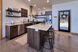 Kitchen with appliances with stainless steel finishes, wall chimney exhaust hood, dark brown cabinetry, sink, and a center island with sink