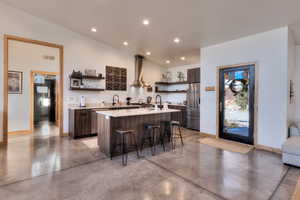 Kitchen featuring wall chimney exhaust hood, a kitchen breakfast bar, an island with sink, dark brown cabinets, and appliances with stainless steel finishes