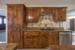 Kitchen featuring stainless steel dishwasher, sink, backsplash, dark stone counters, and light tile patterned floors