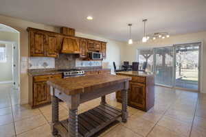 Kitchen featuring custom exhaust hood, appliances with stainless steel finishes, hanging light fixtures, light tile patterned floors, and decorative backsplash