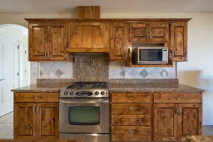 Kitchen with appliances with stainless steel finishes, custom exhaust hood, dark stone countertops, backsplash, and light tile patterned floors