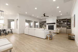 Living room with a stone fireplace, light wood-type flooring, built in shelves, ornamental molding, and ceiling fan