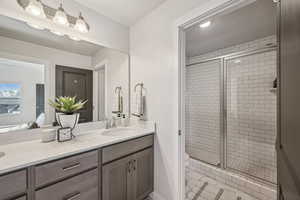 Primary bathroom featuring a tile shower, white quarts countertop with two sinks, a grey wood vanity, and connection to the primary closet