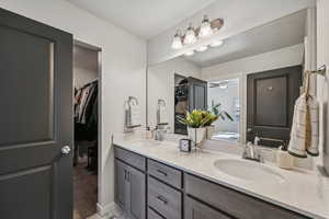 Primary bathroom featuring a tile shower, white quarts countertop with two sinks, a grey wood vanity, and connection to the primary closet