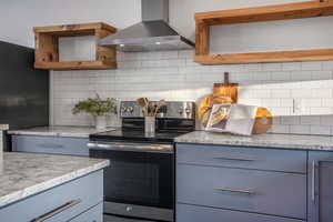 Kitchen with wall chimney range hood, backsplash, blue cabinetry, and stainless steel electric range