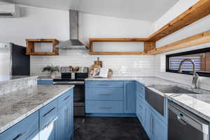 Kitchen with appliances with stainless steel finishes, wall chimney range hood, a wall mounted AC, light stone counters, and blue cabinets