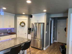 Kitchen featuring stainless steel fridge, hardwood / wood-style flooring, white cabinetry, dark stone counters, and a textured ceiling