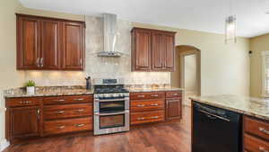 Kitchen featuring light stone countertops, dishwasher, wall chimney range hood, backsplash, and range with two ovens