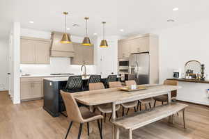 Kitchen with appliances with stainless steel finishes, decorative light fixtures, premium range hood, light wood-type flooring, and an island with sink