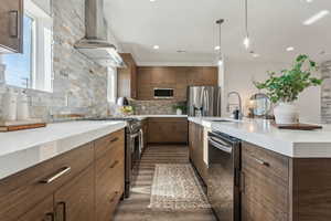 Kitchen with sink, wall chimney range hood, appliances with stainless steel finishes, backsplash, and hanging light fixtures