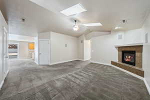 Unfurnished living room with ceiling fan, vaulted ceiling with skylight, a tiled fireplace, and carpet floors