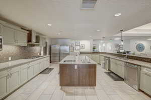 Kitchen with appliances with stainless steel finishes, wall chimney exhaust hood, sink, decorative light fixtures, and a center island with sink