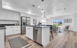 Kitchen featuring sink, appliances with stainless steel finishes, an island with sink, white cabinets, and decorative light fixtures