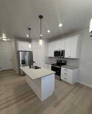 Kitchen featuring sink, stainless steel appliances, an island with sink, white cabinets, and decorative light fixtures