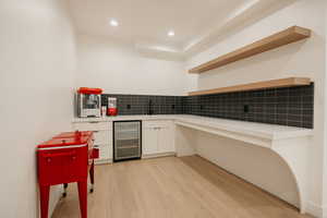 Sink, wine cooler, tasteful backsplash, white cabinets, and light wood-type flooring