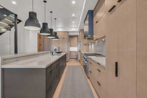 Kitchen with wall chimney range hood, sink, hanging light fixtures, a large island with sink, and light brown cabinets