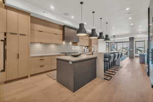 Kitchen featuring light brown cabinetry, hanging light fixtures, a large island, wall chimney range hood, and light wood-type flooring