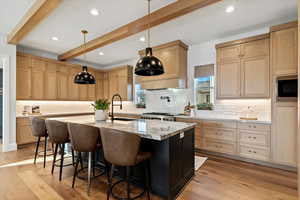 Kitchen with decorative light fixtures, an island with sink, pot filler, 6 burner stove, 2 dishwashers, pebble ice machine in cabinet, light brown cabinets, and beam ceiling