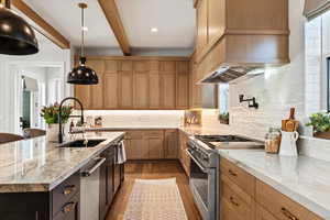 Kitchen with decorative light fixtures, an island with sink, pot filler, 6 burner stove, 2 dishwashers, pebble ice machine in cabinet, light brown cabinets, and beam ceiling